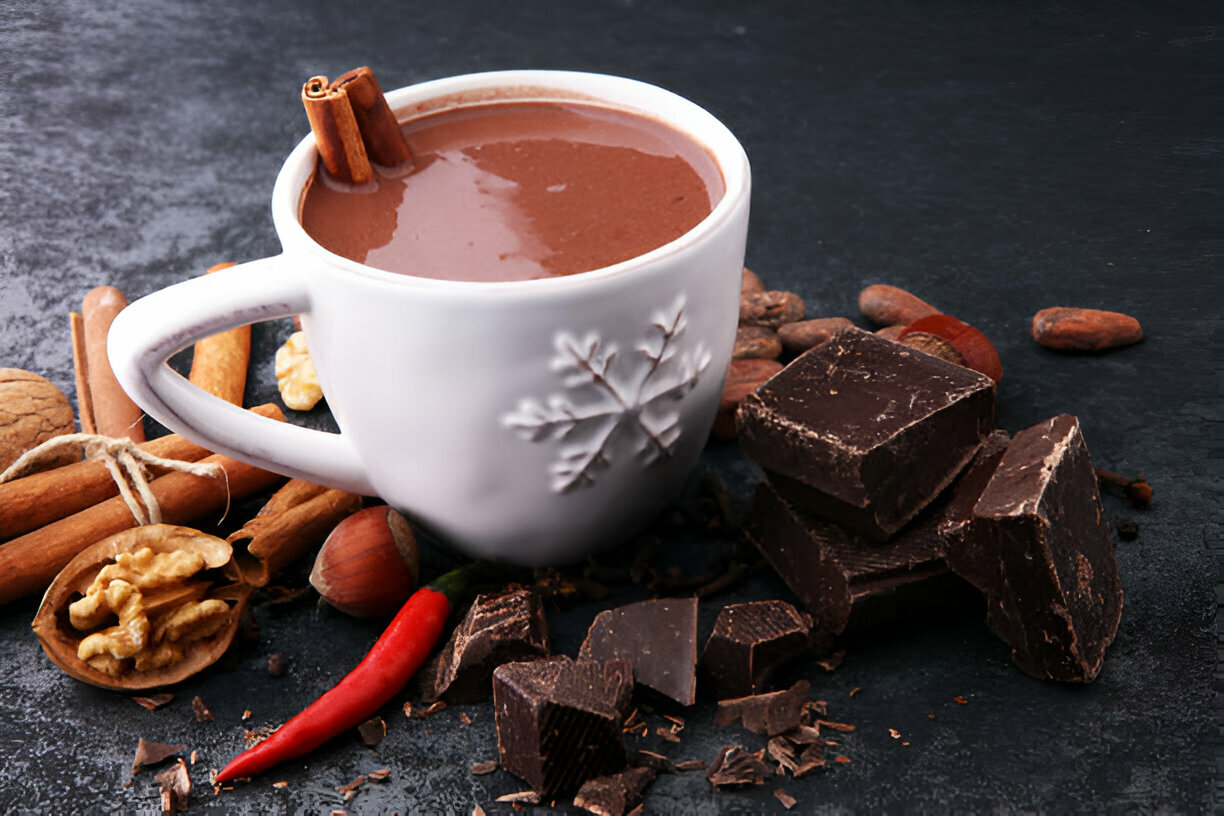 A cozy cup of hot chocolate topped with cinnamon sticks, surrounded by nuts, chocolate pieces, and a red chili pepper.