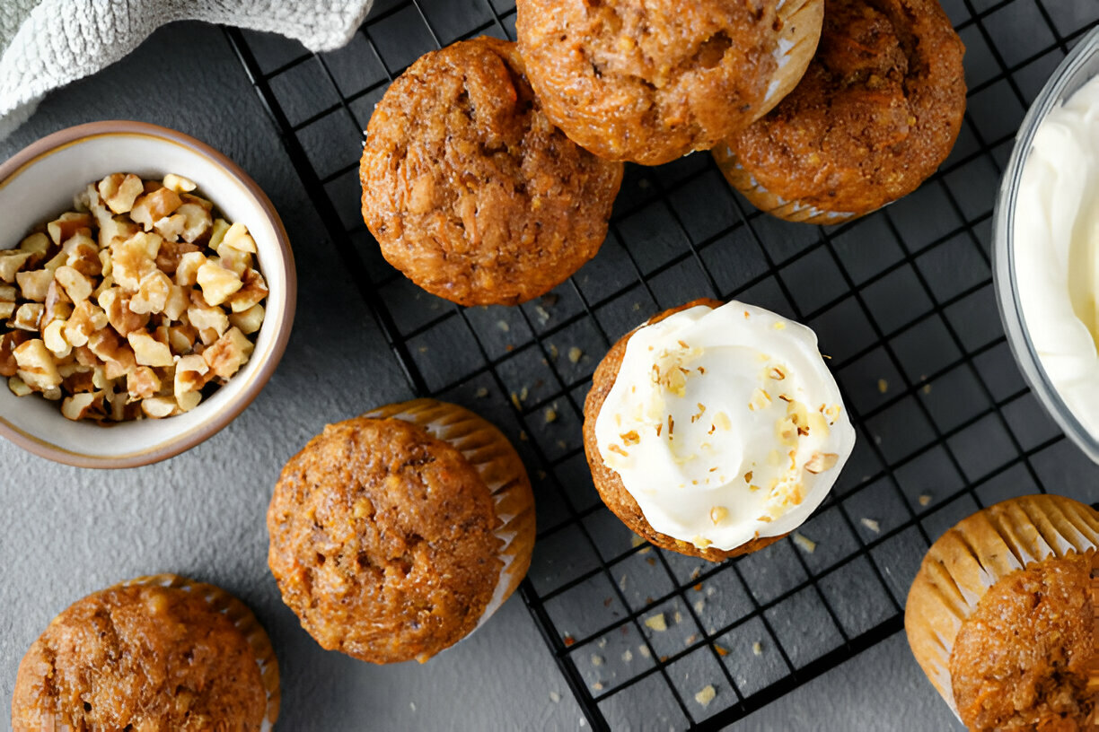 A variety of muffins on a cooling rack, with one frosted muffin and a bowl of chopped walnuts.