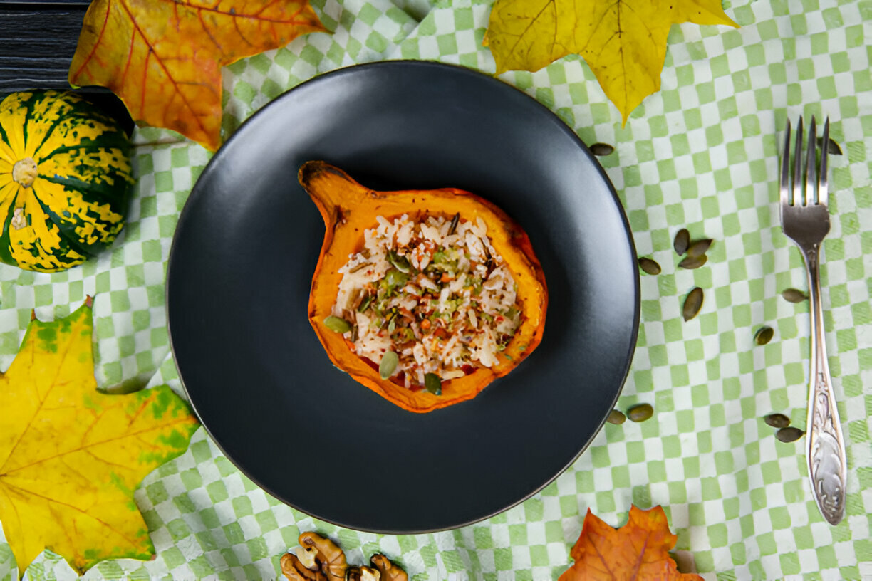 A beautifully presented dish featuring a halved acorn squash filled with rice and vegetables, situated on a dark plate surrounded by fall leaves and decorative gourds.