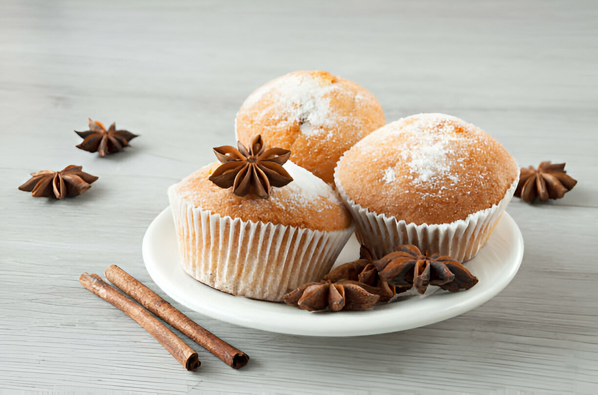 Three freshly baked muffins topped with star anise on a white plate, surrounded by star anise pods and cinnamon sticks.