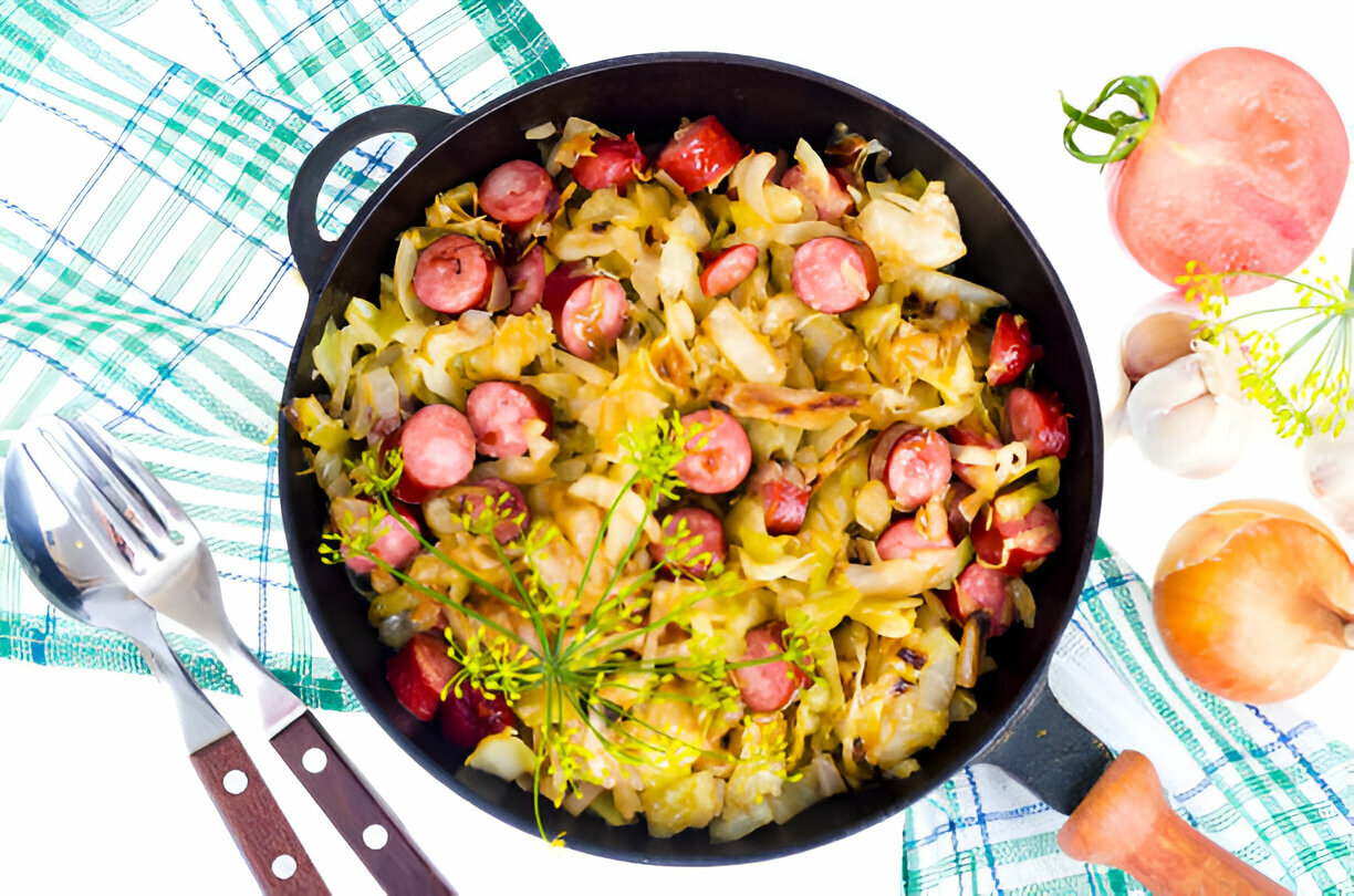 A skillet filled with sautéed cabbage and sliced hot dogs, garnished with fresh herbs, on a checkered cloth.