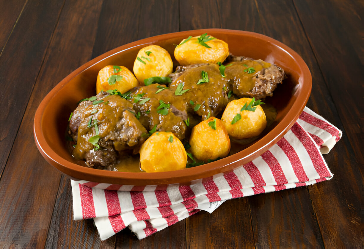 A hearty dish featuring meatloaf with gravy and golden potatoes garnished with fresh herbs, served in a brown ceramic dish on a striped red and white napkin.