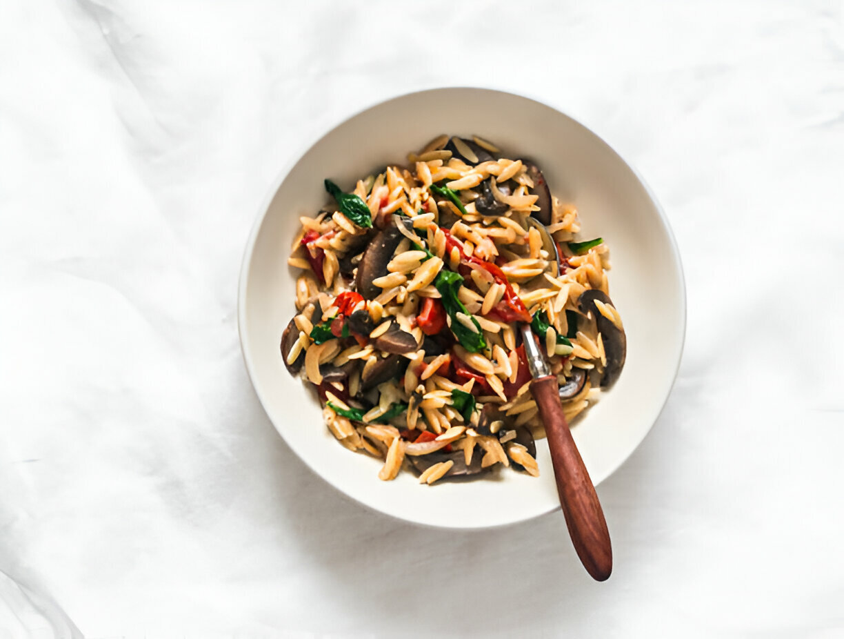 A bowl of orzo pasta mixed with vegetables and herbs, garnished with a wooden spoon.